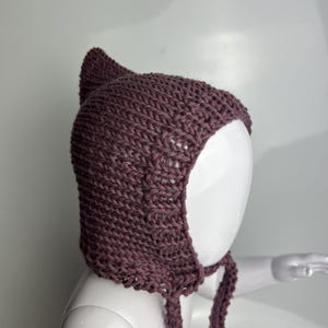 May include: A dark mauve knitted bonnet featuring a pointed top and two long braided ties hanging from the sides. The headwear is crafted with a visible ribbed stitch pattern and covers the crown and sides of the head with a snug fit.