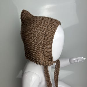 May include: A brown knitted bonnet featuring a pointed top, textured vertical stitch patterns, and two long braided ties hanging from the chin area. The headwear is displayed on a smooth white mannequin head, showcasing the thick yarn construction.