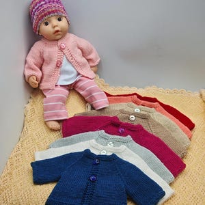 Könnte beinhalten: Eine Puppe trägt einen rosa Strickcardigan, eine Mütze und eine gestreifte Hose. Mehrere andere Strickcardigans in verschiedenen Farben, darunter Blau, Grau und Rosa, werden auf einer strukturierten Oberfläche präsentiert. Die Cardigans haben Knopfverschlüsse.