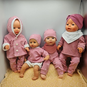 Puede incluir: Cuatro muñecas bebés vestidas con trajes rosas. Las muñecas visten sombreros, camisas, pantalones y chaquetas a juego. Algunos conjuntos tienen detalles de corazones blancos. Una muñeca lleva un gorro tipo turbante y un babero. Las muñecas están posadas sobre una superficie marrón claro.