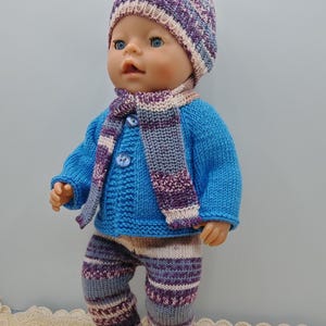 Könnte beinhalten: Ein gestricktes Puppenoutfit in den Farben Lila, Blau und Creme. Das Outfit besteht aus einer Mütze, einem Schal, einem Cardigan und einer Hose.