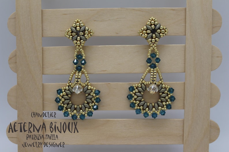 May include: A pair of chandelier earrings with a floral design. The earrings feature gold-coloured beads, teal-coloured crystals, and a central clear crystal. The earrings are displayed on a wooden background. The text "Chandelier" and "Aeterna Bijoux" are visible.