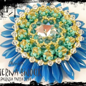 May include: A blue and green beaded brooch with a large crystal in the center. The brooch is surrounded by a ring of smaller beads and has a gold trim. The brooch is labeled "Aeterna Bijoux Patrizia Failla 2014".