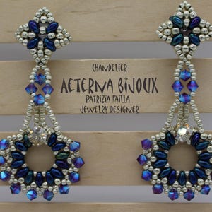 May include: A pair of chandelier earrings with a floral design. The earrings feature silver beads, blue and purple crystals, and a central clear crystal. The text "AETERNA BIJOUX" and "PATRIZIA FAILLA JEWELRY DESIGNER" is visible.