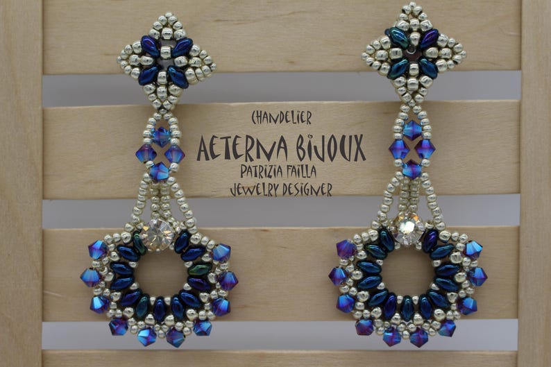May include: A pair of chandelier earrings with a floral design. The earrings feature silver beads, blue and purple crystals, and a central clear crystal. The text "AETERNA BIJOUX" and "PATRIZIA FAILLA JEWELRY DESIGNER" is visible.