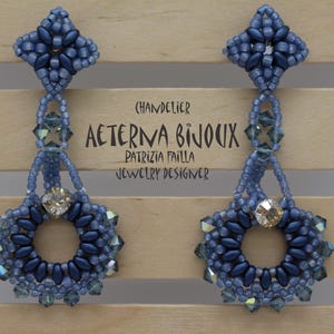 May include: A pair of blue beaded chandelier earrings. The earrings have a floral design with a central crystal and are suspended from a beaded top. The text "AETERNA BIJOUX" is visible. The earrings are displayed against a wooden background.