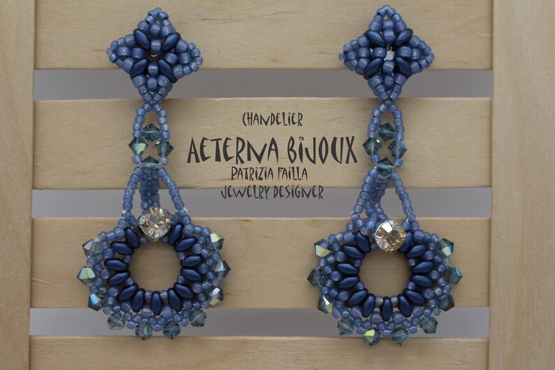 May include: A pair of blue beaded chandelier earrings. The earrings have a floral design with a central crystal and are suspended from a beaded top. The text "AETERNA BIJOUX" is visible. The earrings are displayed against a wooden background.