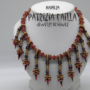 May include: A red, gold, and black beaded necklace with a pointed, dangling design. The necklace is labeled "Nadhera" and "Patrizia Failla Jewelry Designer".