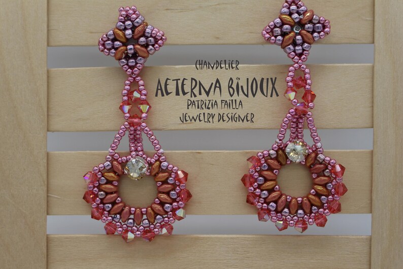 May include: A pair of chandelier earrings in shades of pink, red, and copper. The earrings feature a floral design with a central crystal and are adorned with small beads. The text "AETERNA BIJOUX" is visible.