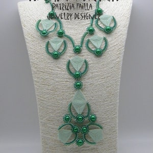 May include: A green and white beaded necklace with a geometric design. The necklace is on a white display stand. The text "PROMETHEUS AETERNA BIJOUX PATRIZIA FAILLA JEWELRY DESIGNER" is visible on the stand.