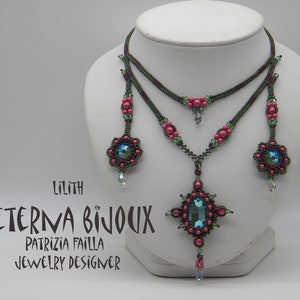 May include: A two-strand beaded necklace with a large, faceted, blue gemstone pendant. The necklace is made with dark green, pink, and iridescent beads. The pendant is surrounded by smaller pink beads. The necklace is displayed on a white mannequin. The text "Lilith Aeterna Bijoux Patrizia Failla Jewelry Designer" is printed below the necklace.