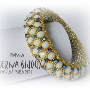 May include: A white and gold beaded bracelet with blue iridescent beads. The bracelet is on a white surface with the text "DHARMA AETERNA BIJOUX PATRIZIA FAILLA 2014".