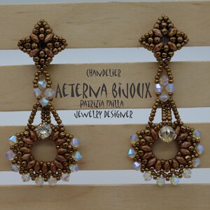 May include: A pair of chandelier earrings in a bronze beaded design. The earrings have a floral top, a beaded chain, and a circular bottom with a central crystal. The text "AETERNA BIJOUX" is visible. The earrings are a statement piece.