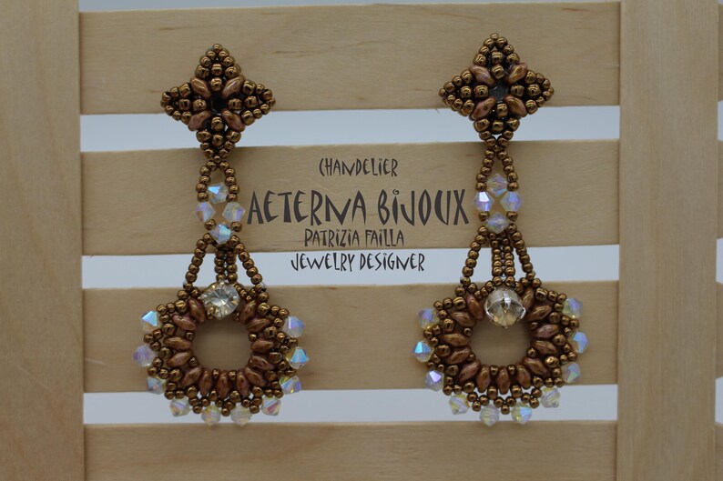 May include: A pair of chandelier earrings in a bronze beaded design. The earrings have a floral top, a beaded chain, and a circular bottom with a central crystal. The text "AETERNA BIJOUX" is visible. The earrings are a statement piece.