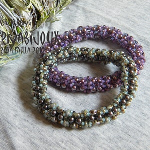 May include: Two beaded bracelets, one with purple and light blue beads, the other with brown and light blue beads. The bracelets are twisted together and are on a gray fabric background.