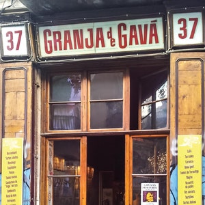 May include: A shop front with a sign that reads "Granja de Gavà" in red letters. The shop front has a wooden door and windows with a chalkboard menu that reads "Iberic Cold Cuts 'n Cheese, Salads 'n Pizza, Wine Menu, Vermut Homemade, Sangria, Beer Cava, Mojito..." and another chalkboard menu that reads "Deliciosas Torrades, Talles d'Embotits i Formatges, Amandes Pizza, Creps Gofres, Aperitius Espinaler, Carta de Vins, Vermut Cava, Modito Caipirinha".