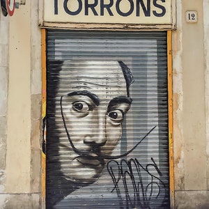 May include: A black and white graffiti mural of Salvador Dali's face on a metal roller shutter. The door is labelled "TORRONS" in black letters on a yellow sign above the door. The number "12" is on the wall to the right of the door.