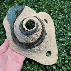 May include: Handmade ceramic art piece with a beige base and teal and brown accents. The piece features a central circular design with a hole, surrounded by textured patterns and several smaller holes. The artwork is held against a green background.