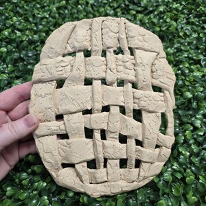 May include: A beige, oval-shaped ceramic piece with a woven basket design. The ceramic has a textured surface and is held against a backdrop of green foliage. The basket weave pattern is detailed and raised.