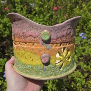 May include: Handmade ceramic planter with a vibrant color gradient from green to yellow, orange, and pink. The planter features decorative cutouts and embossed patterns. Two green and pink knobs add detail to the design.