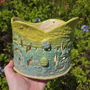 May include: A handmade ceramic planter with a textured, layered design. The planter features a vibrant color palette of yellow, green, and blue, with cut-out leaf shapes. Two decorative, round buttons are on the side. The planter is held by a hand.
