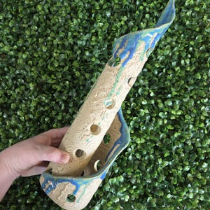 May include: A cylindrical ceramic flute with a textured, speckled beige surface featuring embossed circular patterns. The instrument has multiple finger holes and is accented with a vibrant blue glaze that drips down the sides and top.