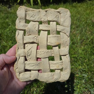 May include: A beige ceramic woven wall hanging with textured details. The rectangular piece features an interwoven design with embossed patterns. The artwork is held in a hand against a backdrop of green grass.