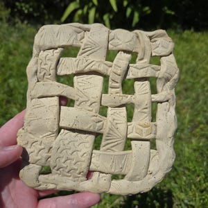 May include: A beige ceramic piece with a woven design. The ceramic has a textured surface and is decorated with various geometric patterns. The piece is held in a hand against a background of green grass.