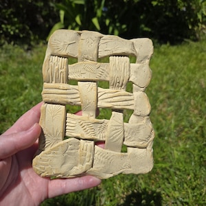 May include: A beige ceramic woven wall hanging with embossed botanical designs. The rectangular piece features a basket weave pattern with detailed impressions of leaves and other natural elements. The handmade art piece is held in a hand against a green background.