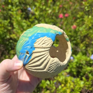 May include: A hand-held, spherical ceramic pot with a large opening. The pot is beige with blue and green painted accents. The surface has carved, fan-like patterns. The background is blurred green foliage.