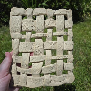 May include: A beige ceramic woven wall hanging with embossed floral and geometric patterns. The rectangular piece has a textured surface and is held by a hand against a backdrop of green grass. The design resembles a basket weave.