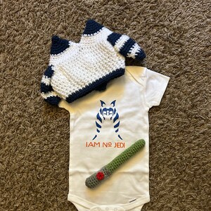 May include: A white baby onesie with a blue and white image of a Star Wars character and the text "I AM NO JEDI". A green crocheted lightsaber and a white crocheted hat with black stripes are also included.