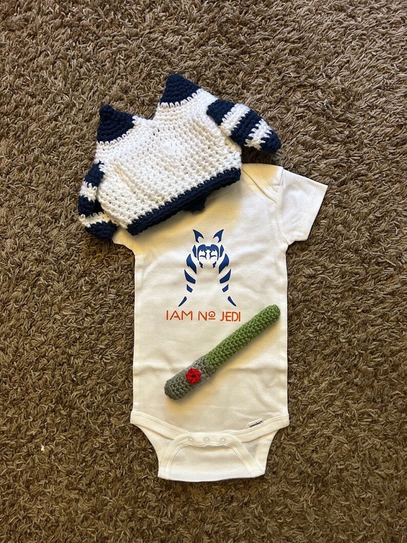 May include: A white baby onesie with a blue and white image of a Star Wars character and the text "I AM NO JEDI". A green crocheted lightsaber and a white crocheted hat with black stripes are also included.