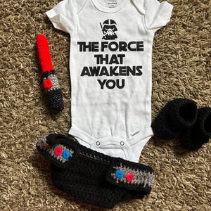 Op de afbeelding: Een witte baby-onesie met een Darth Vader-afbeelding en de tekst "THE FORCE THAT AWAKENS YOU". De set bevat een gehaakte lichtzwaard, een luier met rode en blauwe knopen en zwarte babyschoentjes.