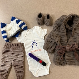 Op de afbeelding: Een baby-outfitset met een wit rompertje met de tekst "I AM NO JEDI" en een zwart-wit gestreepte afbeelding van een personage uit Star Wars. De set bevat ook een bruine gebreide gekapte jas, een bruine gebreide broek, een wit-blauwe gebreide muts en een paar bruine gebreide slofjes.