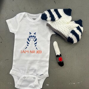 May include: White baby onesie with a black and white striped character and the text "I AM NO JEDI". A white and blue striped crocheted hat and a white crocheted finger puppet with a red circle on the end.