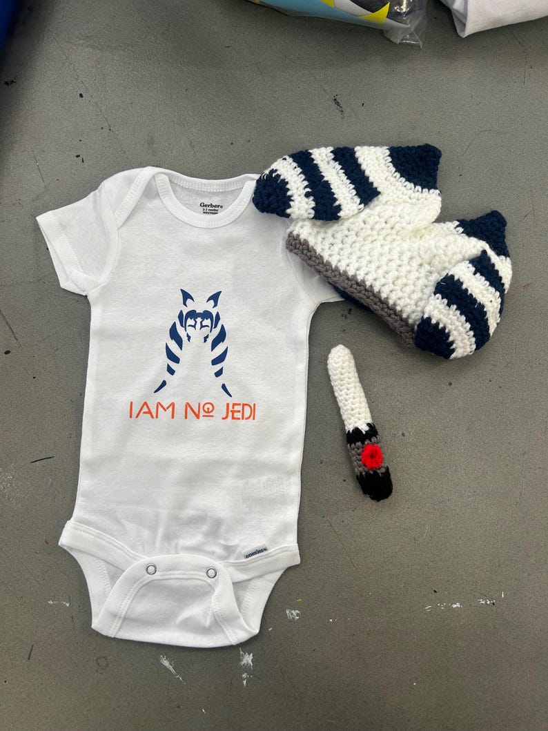 May include: White baby onesie with a black and white striped character and the text "I AM NO JEDI". A white and blue striped crocheted hat and a white crocheted finger puppet with a red circle on the end.