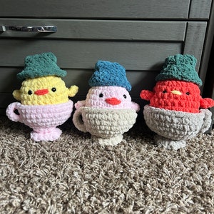 Puede incluir: Tres animales de peluche de crochet con forma de pájaros sentados en tazas de té. Los pájaros llevan sombreros y tienen cuerpos de diferentes colores: amarillo, rosa y rojo.