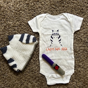 May include: White baby onesie with a black and white image of a Star Wars character and the text "I AM NO JEDI" in orange. A purple and gray crocheted lightsaber and a white and black striped crocheted hat are also pictured.