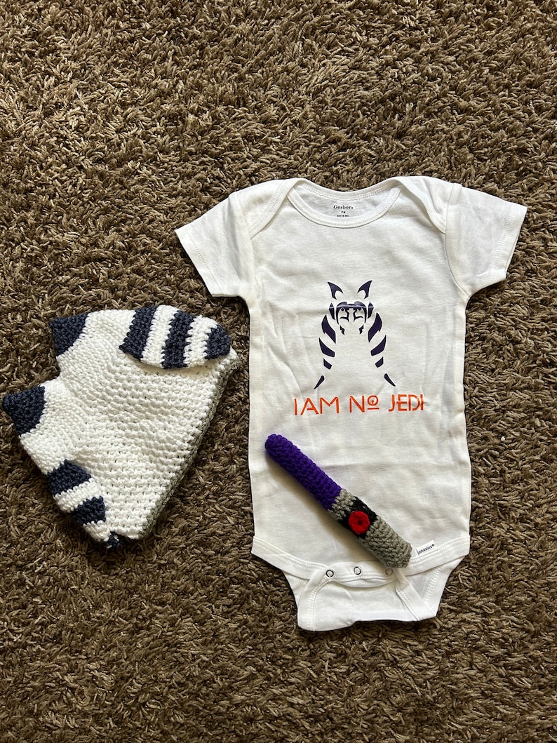 May include: White baby onesie with a black and white image of a Star Wars character and the text "I AM NO JEDI" in orange. A purple and gray crocheted lightsaber and a white and black striped crocheted hat are also pictured.