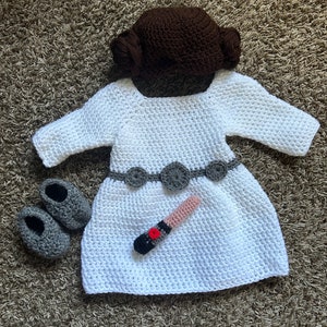 Op de afbeelding: Een wit gebreid babyjurkje met een bruin gebreide muts, een grijze gebreide riem en een roze lightsaber. Het jurkje heeft een ronde hals en lange mouwen. De muts heeft twee knotjes bovenop. De lightsaber ligt op het jurkje.