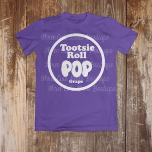 May include: A purple t-shirt with a white circle graphic that says 'Tootsie Roll POP Grape'.