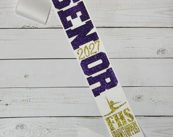 Puede incluir: Una banda de satén blanco con texto brillante en dorado y morado. La banda muestra el nombre Lora, la palabra SENIOR, el año 2027, una silueta de gimnasta, las letras FHS y la palabra SHOWSTOPPERS en una disposición vertical.