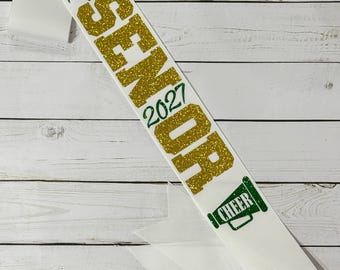 Puede incluir: Una banda blanca con el nombre Taylor en letra cursiva verde, la palabra SENIOR en letras grandes de purpurina dorada, el año 2027 en verde y un icono de megáfono verde con la palabra CHEER impresa en su interior. La banda es de tela lisa.
