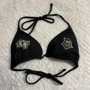 May include: Black bikini top with UCF logo and a knight emblem. The top has adjustable ties at the neck and back. The UCF logo is in gold and white. The knight emblem is in gold, black, and white.