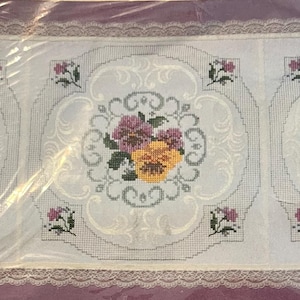 May include: A white embroidered table runner featuring a floral design. The runner has three central panels with a large pansy in the center, surrounded by a decorative border. The edges are trimmed with lace.