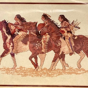 Puede incluir: Obra de arte de punto de cruz que representa a tres figuras nativas americanas montadas a caballo. Los caballos son marrones y blancos, y los jinetes visten ropa tradicional. La obra está enmarcada y sobre un fondo crema.