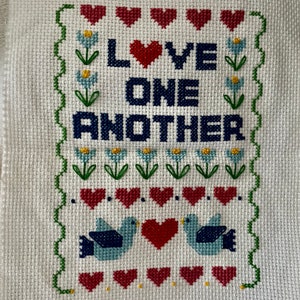 May include: Cross-stitch embroidery on white fabric. The design features the words "LOVE ONE ANOTHER" in navy blue, with a red heart replacing the "O" in "LOVE". The design includes red hearts, blue birds, and flowers.