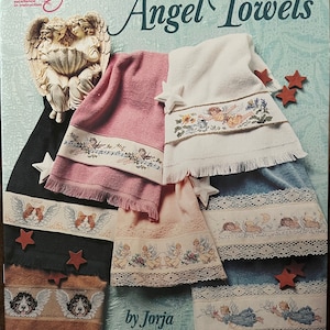 Puede incluir: Un libro de patrones de punto de cruz titulado "Angel Towels" de Jorja Hernandez. La portada presenta varias toallas con dise&ntilde;os de punto de cruz con tem&aacute;tica de &aacute;ngeles, incluyendo &aacute;ngeles, patrones florales y caras de perros. Las toallas son en tonos rosa, blanco, azul y beige.