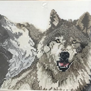 May include: A cross-stitch artwork featuring four wolves in varying shades of gray and black. The wolves are depicted in a row, with their heads and upper bodies visible. The central wolf has its mouth open, revealing a pink tongue.